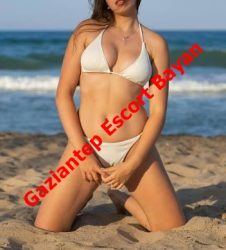 Woman in a white bikini kneels in sand at the beach with the sea in the background; red watermark across reads 'Gaziantep Escort Bayan'.