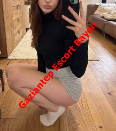 Mirror selfie of a woman in a black turtleneck and gray shorts, crouching on a wooden floor; red diagonal text 'Gaziantep Escort Bayan' across.