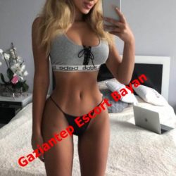 Woman posing in a gray sports bra and black thong taking a mirror selfie in a bedroom; red banner text reads 'Gaziantep Escort Bayan' across her torso.